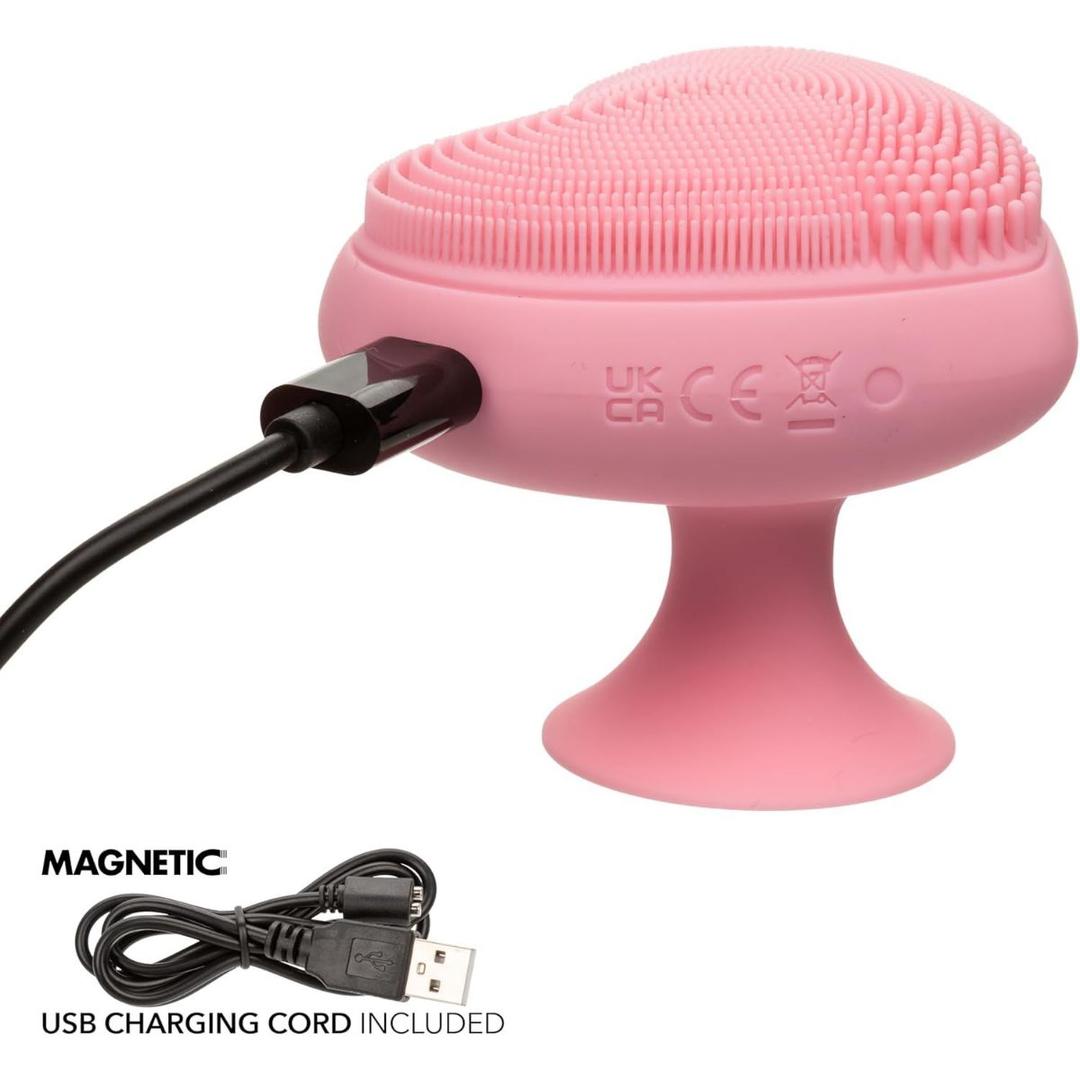 The CalExotics Posh Kiss Me Handheld Massager, featuring a pink heart-shaped design with textured pleasure nubbies and a suction base, is shown charging via a magnetic USB cable. The charging cord appears separately below the device.