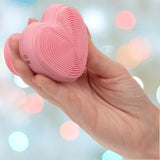 A hand holds the CalExotics Posh Kiss Me Handheld Massager with textured pleasure nubbies and a suction base against a blurred background of pastel-colored lights.