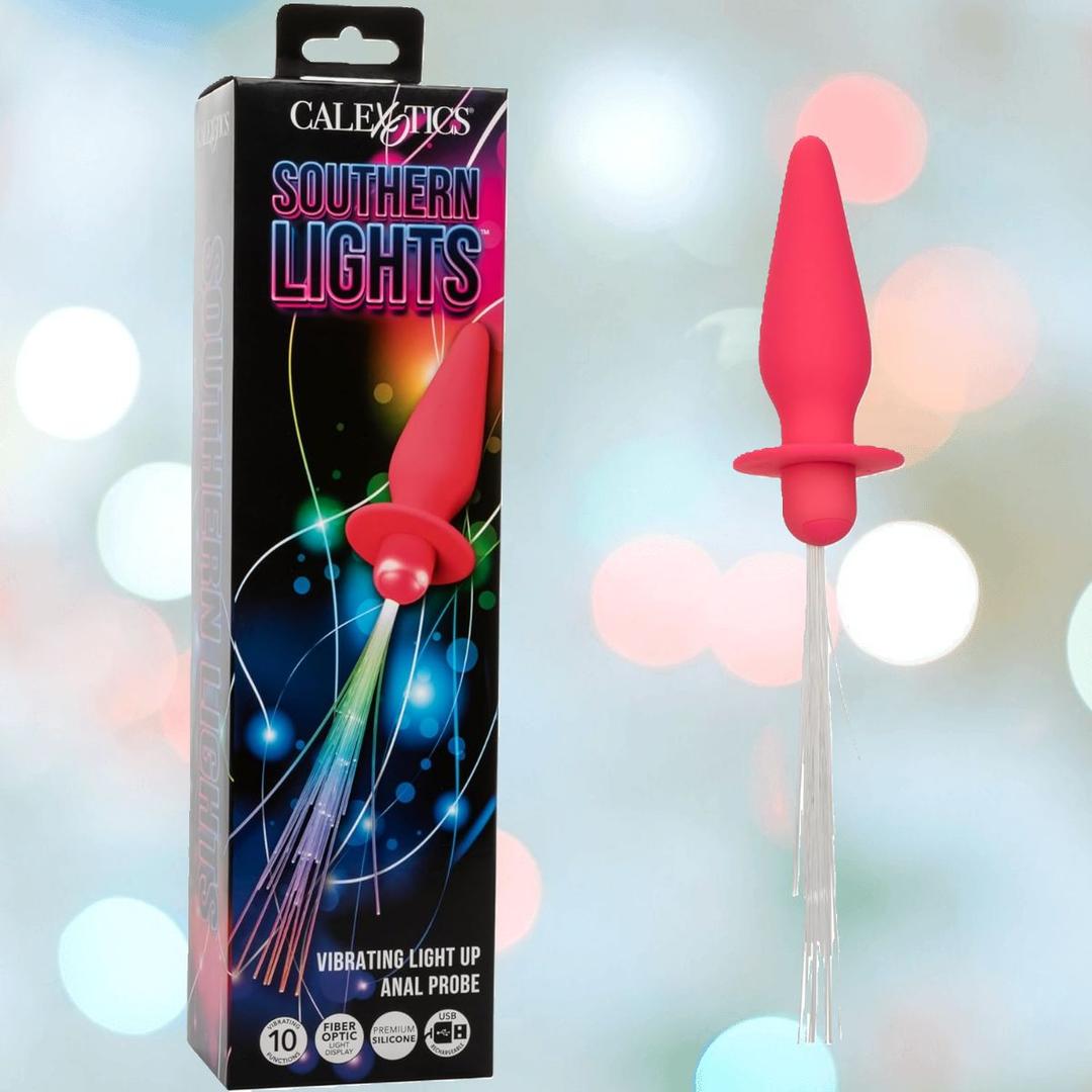 A pink CalExotics Southern Lights Vibrating Light Up Butt Plug Anal Vibrator Probe is displayed beside its vibrant box, set against a blurred pastel background.