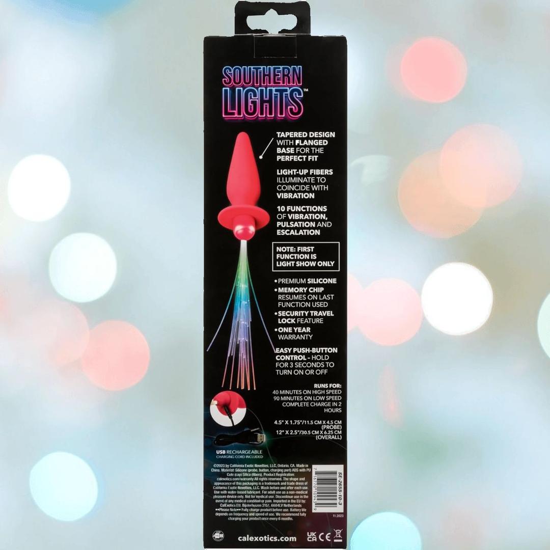 The CalExotics Southern Lights Vibrating Light Up Butt Plug Anal Vibrator Probe – Pink features a tapered, pink rechargeable silicone design with multi-colored LED lights, vibration settings, USB charging, and remote control on a soft bokeh-themed box.