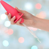 A hand holds the CalExotics Southern Lights Vibrating Light Up Butt Plug Anal Vibrator Probe - Pink, with water streaming from its nozzle against a pastel-colored, blurred background.