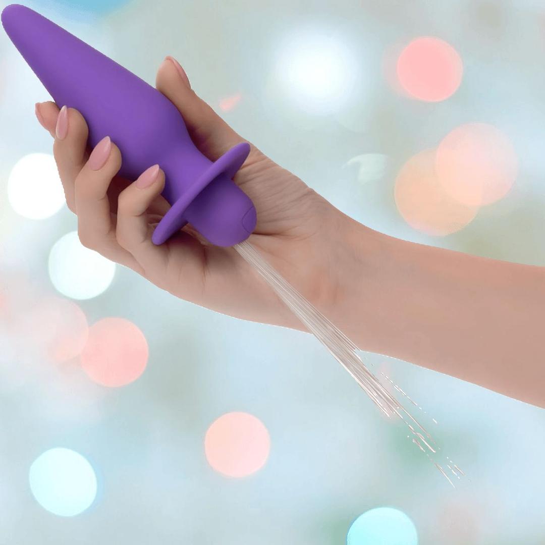A hand holds the CalExotics Southern Lights Vibrating Light Up Butt Plug Anal Vibrator Probe in purple, with water spraying from its tip, set against a backdrop of blurred colorful lights.