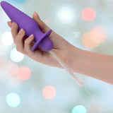 A hand holds the CalExotics Southern Lights Vibrating Light Up Butt Plug Anal Vibrator Probe in purple, with water spraying from its tip, set against a backdrop of blurred colorful lights.