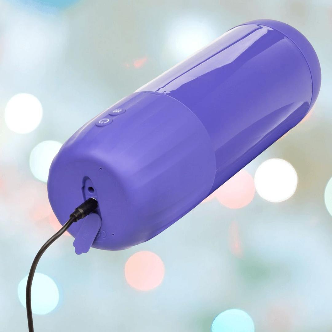 A close-up of the CalExotics Connect Stroker with Vibration and Suction, a purple cylindrical device by CalExotics, being charged with a black cable against a softly blurred background featuring circular light spots.