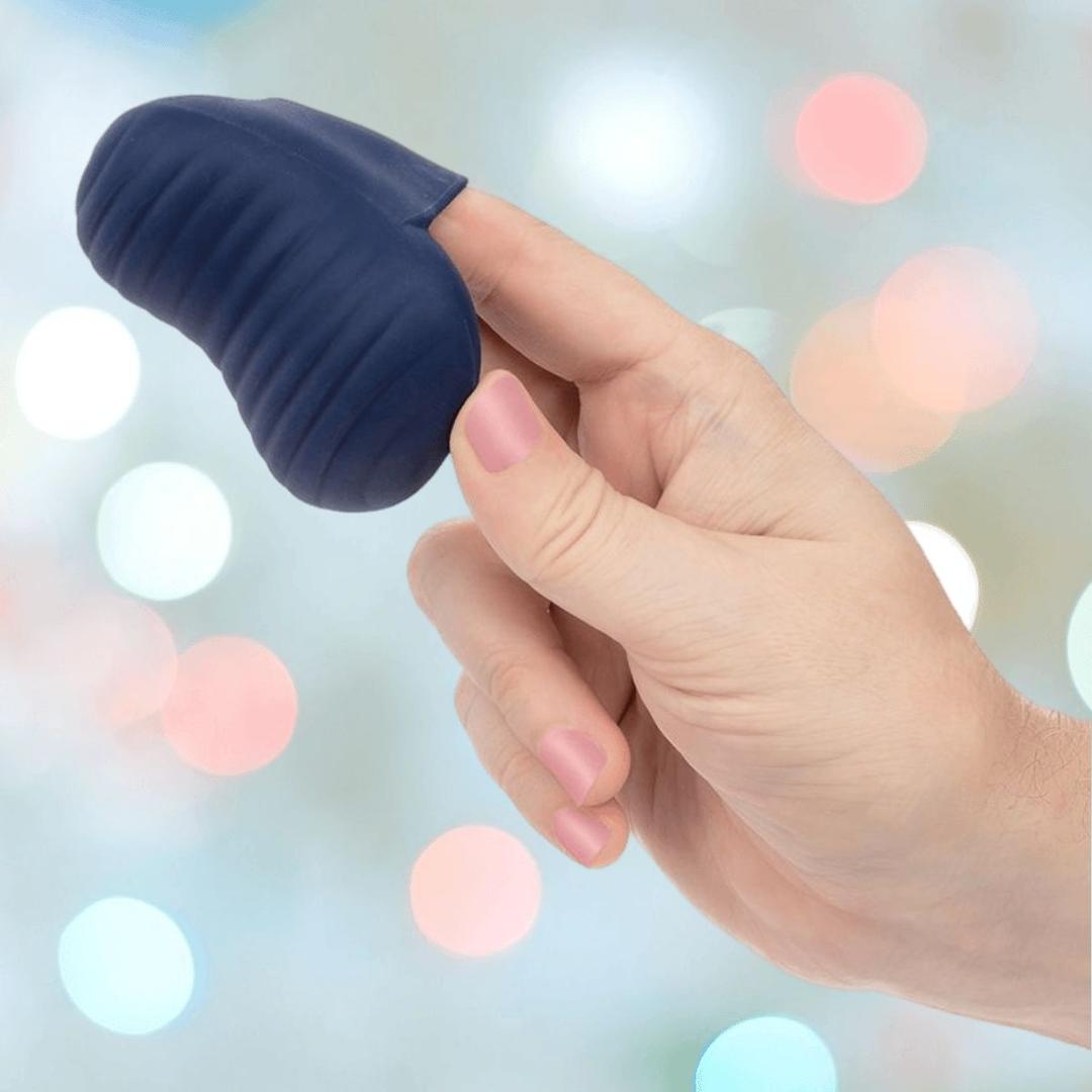 A hand with pink-painted nails holds the CalExotics Cashmere Dual Finger Massager, a small navy blue ridged silicone device, on the index and middle fingers against a softly blurred pastel bokeh background.