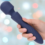 A hand holds the CalExotics Charisma Mystique Wand Massager with flexible head, featuring two side buttons and a dark blue design, set against a blurred pastel bokeh background.