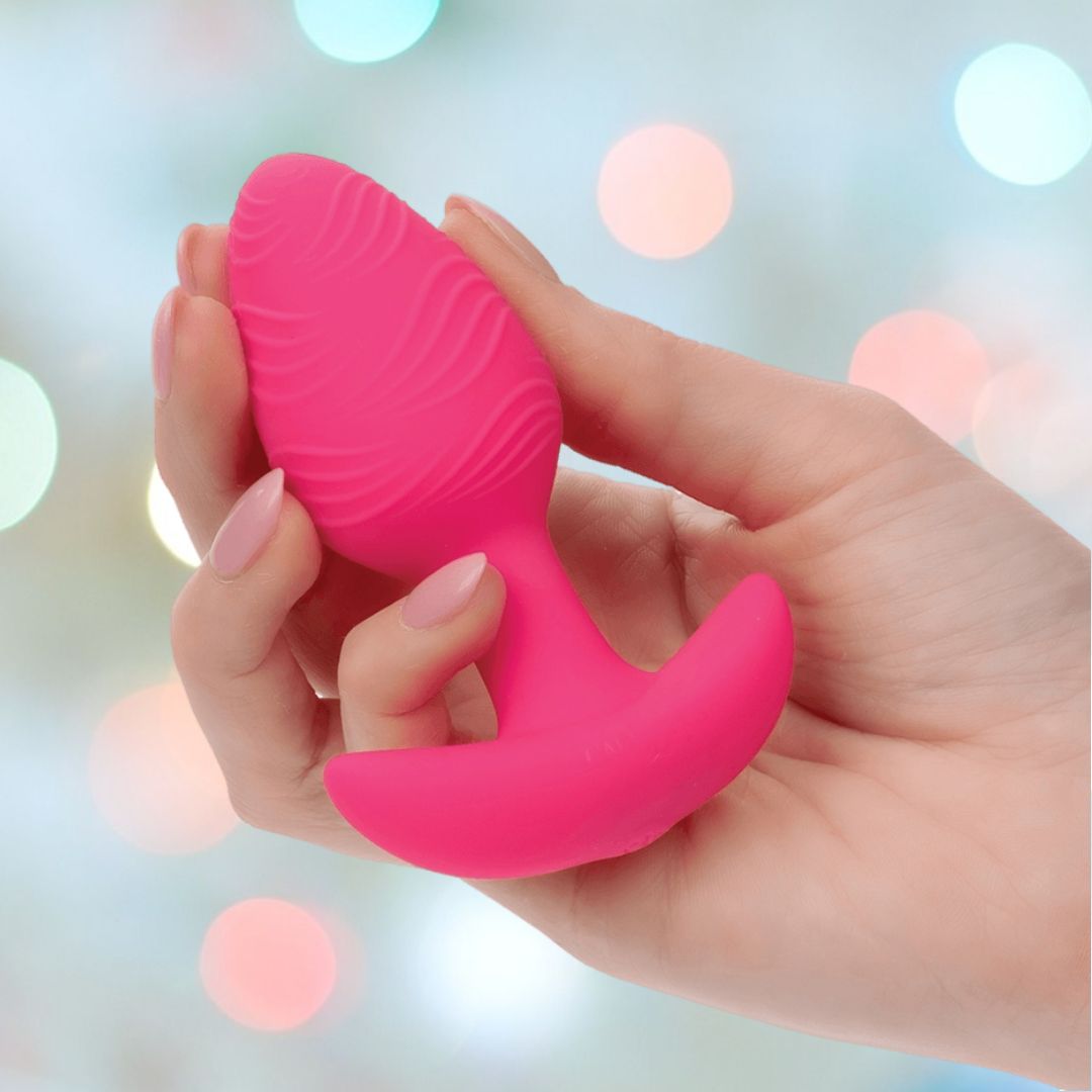 A hand displays the pink, textured Cheeky Vibrating Glow-In-The-Dark Butt Plug by CalExotics, crafted from body-safe silicone with a curved base, set against a blurred backdrop of colorful bokeh lights.