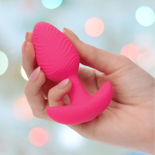 A hand displays the pink, textured Cheeky Vibrating Glow-In-The-Dark Butt Plug by CalExotics, crafted from body-safe silicone with a curved base, set against a blurred backdrop of colorful bokeh lights. 1080