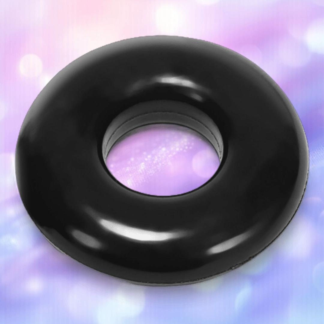 A best-selling Oxballs Atomic Jock "Do-Nut-2" Large cock ring, sleek and black with a donut shape, is centered against a softly blurred pastel background in pink, purple, and blue tones.