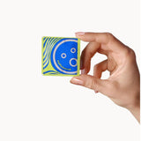 A hand with light skin holds a blue and green packet of Dame Ultra-Thin Vegan Friendly Latex Condoms, featuring a playful smiley face design, against a plain white background.