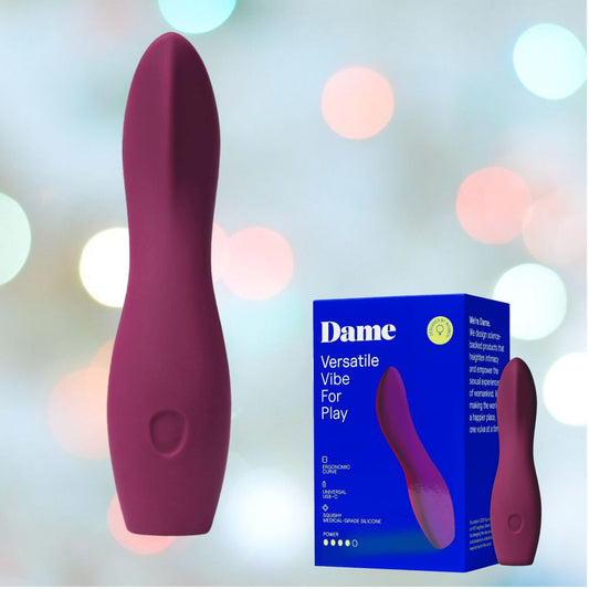 A Plum-colored Dame Products bullet vibrator, known as the Dame Dip 2.0, sits beside its blue packaging box labeled "Dame Versatile Vibe For Play." The backdrop is adorned with softly blurred pastel lights, highlighting its ergonomic design. This vibrator is USB-C rechargeable, offering both convenience and modern functionality. 1080