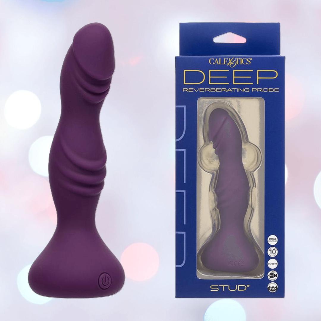 A purple, body-safe silicone probe with a rippled, textured curve stands by its blue and white CalExotics packaging labeled “Deep Stud Reverberating Probe: Rippled Textured Vibrator” against a softly blurred pastel background.