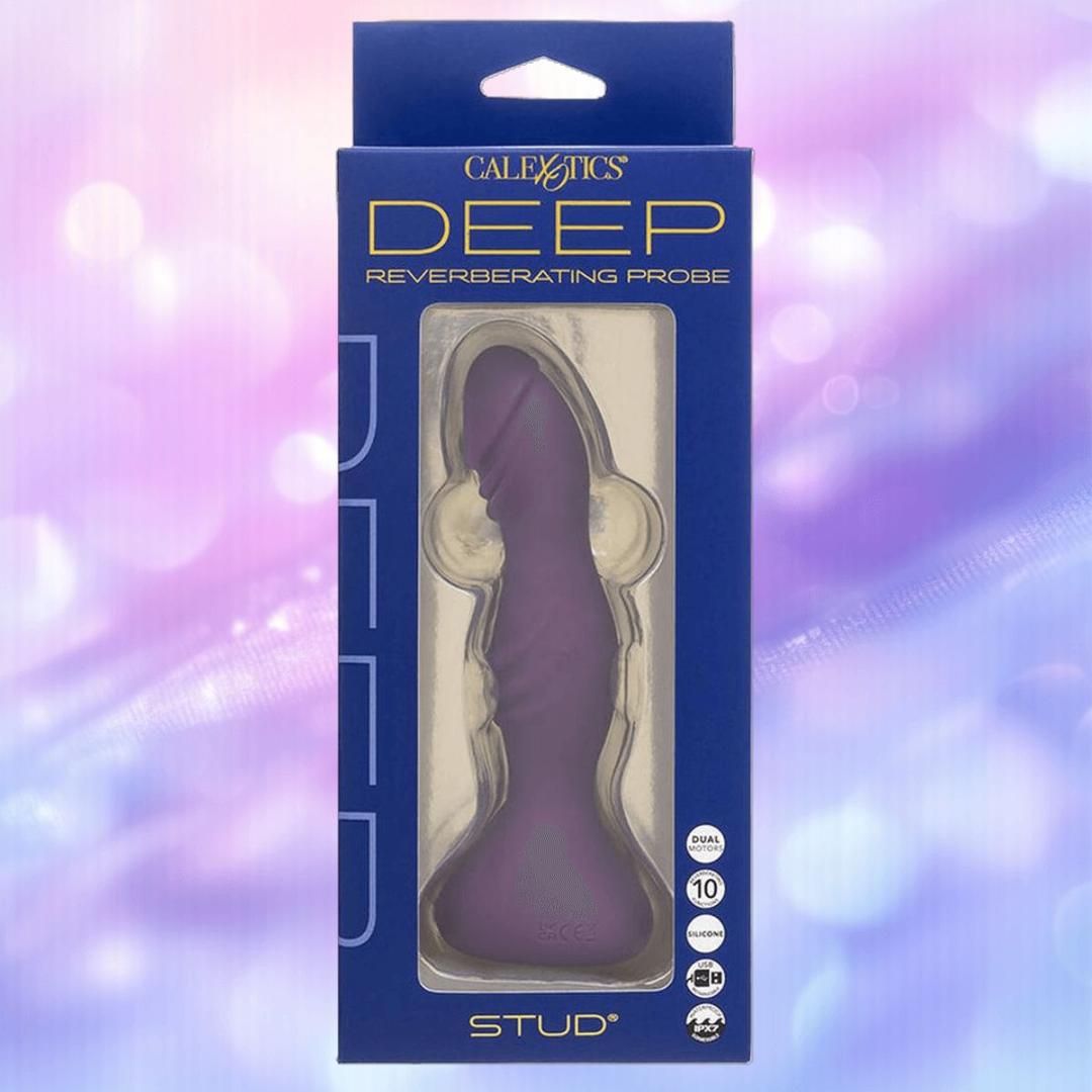 A boxed CalExotics Deep Stud Vibrating Probe in purple, made of body-safe silicone, is shown against a pastel purple and pink blurred background. The packaging displays features and the product name "STUD" at the bottom.
