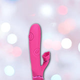 The Dolce Vive Fiore Rechargeable G-Spot Air Wave Vibrating and Tapping Clit by Shots America, a pink silicone vibrator with curved shaft and rose-shaped clitoral stimulator, is displayed against a soft, blurred pastel background.