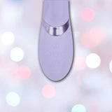 A close-up of the lavender Dolce Vive Fronda Rechargeable G-Spot Vibrating and Tapping Leaf Clit Stim by Shots America, featuring four buttons and the "VIVE" logo, set against a pastel bokeh background of pink, blue, and white lights.