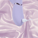 The Dolce Vive Fronda Rechargeable G-Spot Vibrating and Tapping Leaf Clit Stim by Shots America, in lavender, is charging with a white cable on a backdrop of light pink, silky fabric.