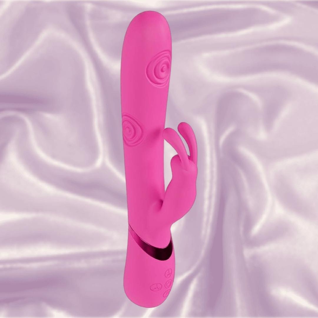 The Shots America Dolce Vive Mimi Rechargeable G-Spot Vibrating and Tapping Rabbit in pink, featuring dual stimulation arms and a spiral texture, stands upright on soft light purple satin fabric.