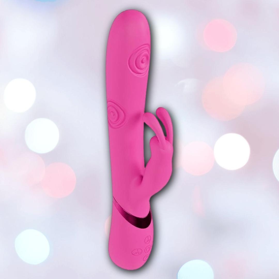 The Shots America Dolce Vive Mimi Rechargeable G-Spot Vibrating and Tapping Rabbit-Pink features a main shaft and clitoral stimulator with spiral textures and base controls, shown against a blurred pastel background.