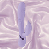 The Dolce Vive Pallina Rechargeable G-Spot Vibrating and Tapping Beads Clit by Shots America is a lavender vibrator with a smooth, curved design and clit stimulator, shown on silky light purple fabric.