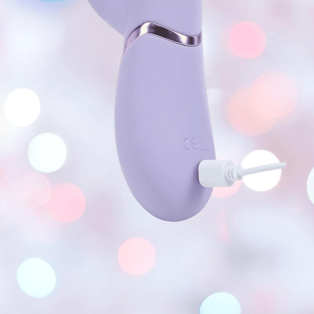 A close-up of the lavender Dolce Vive Fronda Rechargeable G-Spot Vibrating and Tapping Leaf Clit Stim from Shots America, charging with a white cable against a softly blurred pastel background with pink and white lights.
