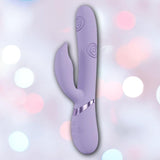 The Shots America Dolce Vive Pallina is a lavender rechargeable G-spot and clit vibrator with a curved shaft, textured spiral patterns, silver accents, and set against a soft pastel bokeh background.