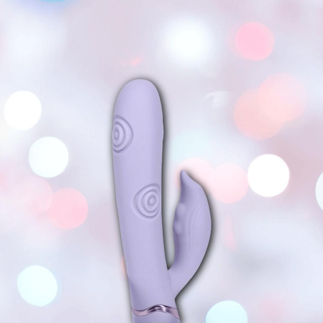 The Dolce Vive Pallina Rechargeable G-Spot Vibrating and Tapping Beads Clit by Shots America is shown in lavender with a curved shaft and external stimulator, set against a soft, pastel bokeh background.