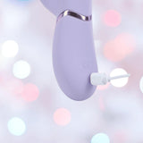 A close-up of the Dolce Vive Pallina Rechargeable G-Spot Vibrating and Tapping Beads Clit by Shots America being charged with a white cable, set against a soft background of pink and white blurred lights.