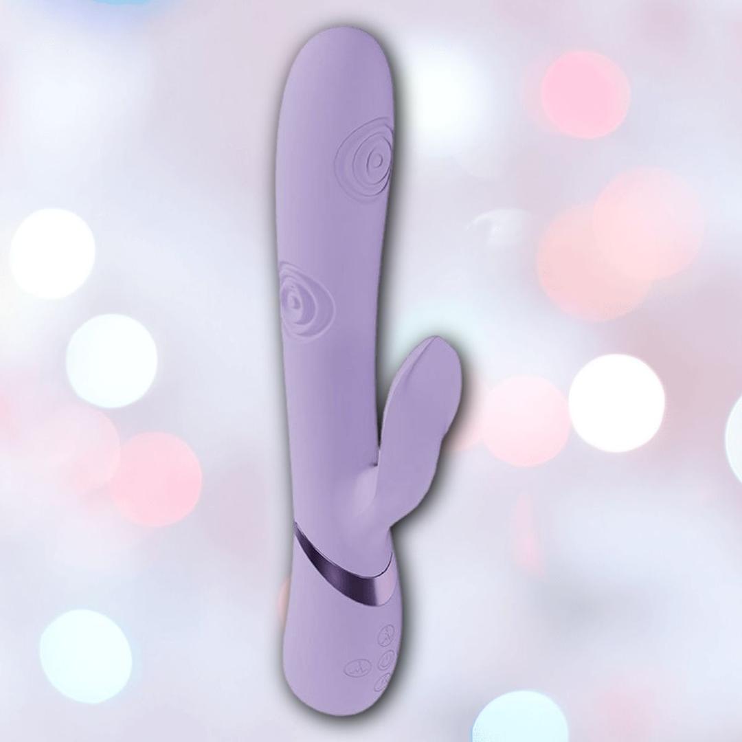 The Dolce Vive Pallina Rechargeable G-Spot Vibrating and Tapping Beads Clit by Shots America features a lavender silicone curved shaft and smaller external stimulator, shown against a blurred pastel bokeh background.