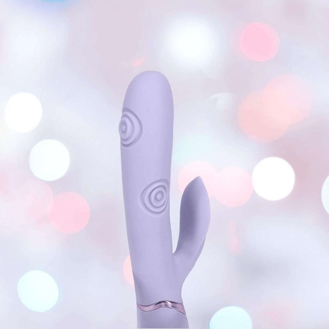 The Dolce Vive Pallina by Shots America is a lavender rechargeable G-spot toy with a curved, textured shaft and smaller clit attachment, shown on a soft pastel bokeh background.