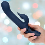A hand holds the Orgasmatron Digital G-Teaser by CalExotics—a dark blue silicone vibrator with a curved tip and a smaller extension—set against a blurred backdrop of pastel lights.
