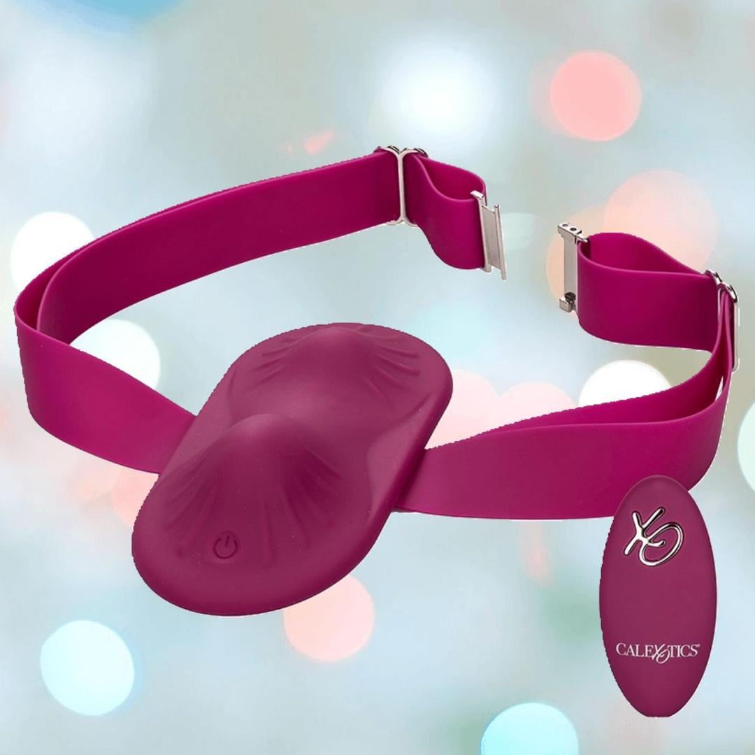The CalExotics Dual Rider Remote Control Grinder is a magenta wearable strap-on vibrator with dual molded shapes, adjustable strap, and matching wireless remote, shown on a soft background with pastel bokeh lights.