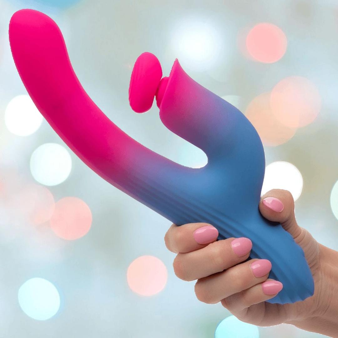 A hand with pink nails holds the CalExotics Elation Thrusting Dual Stimulator Rabbit Vibrator, a blue and pink silicone toy with dual tips and curved design, set against a softly blurred pastel bokeh background.
