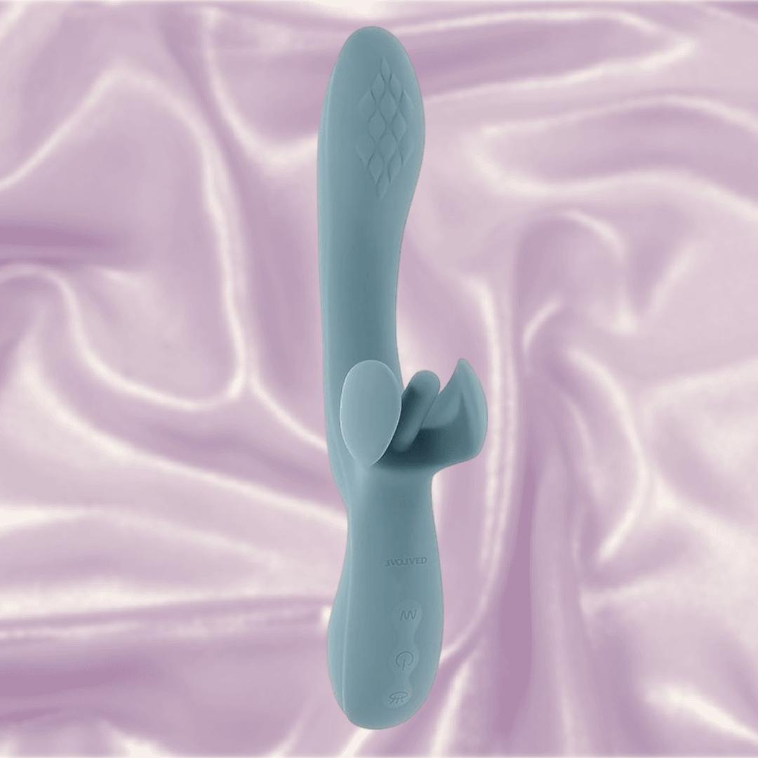 The Evolved Novelties Evolved Take Your Flick is a teal silicone dual stimulation vibrator with textured detail and an external stimulator, shown upright on light purple fabric. Enjoy waterproof, customizable pleasure for intimate moments.