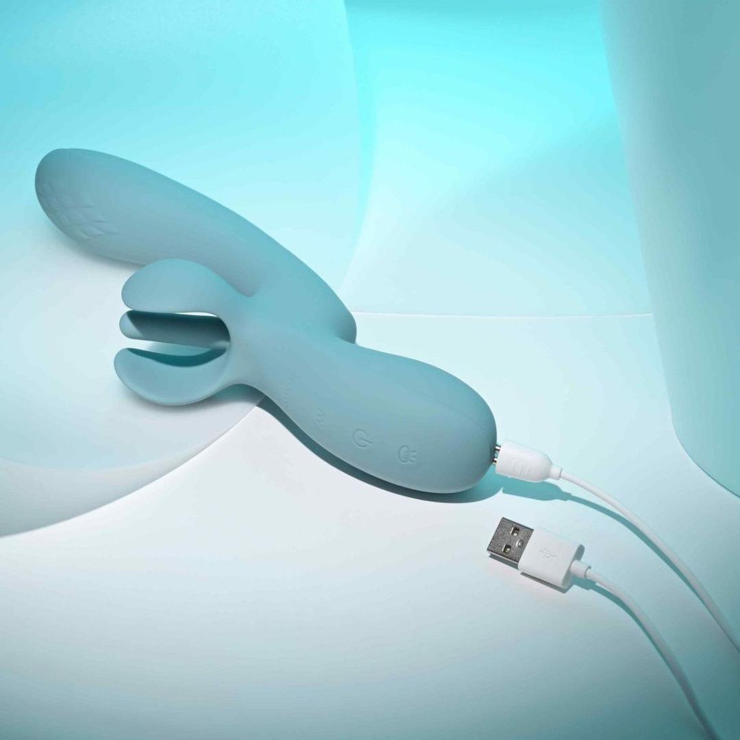 The Evolved Take Your Flick, a blue silicone flickering vibrator by Evolved Novelties with a split tip, charges via a white USB cable against a soft blue and white background with gentle abstract lighting, promising customizable pleasure.