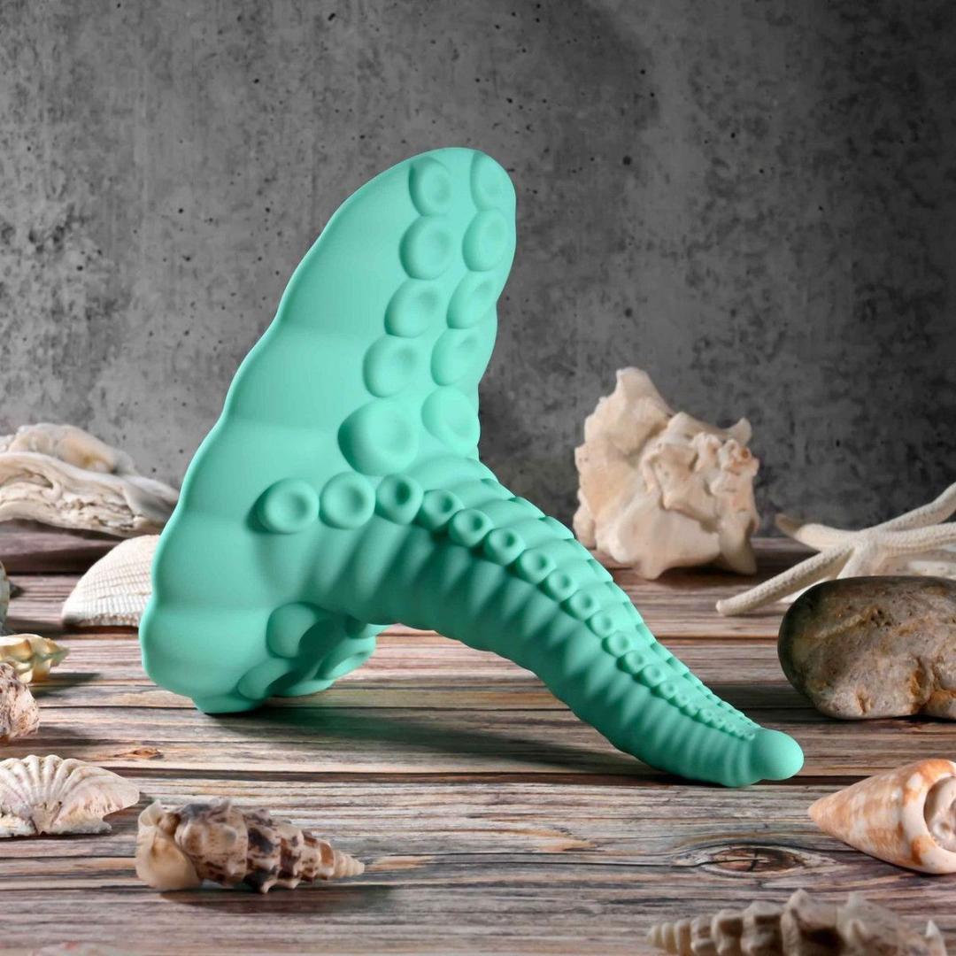 The Evolved Tentacle Grinder by Evolved Novelties, shaped like a teal octopus tentacle with realistic suction cups, stands upright on a wooden surface surrounded by seashells and coral against a textured gray background.