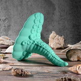 The Evolved Tentacle Grinder by Evolved Novelties, shaped like a teal octopus tentacle with realistic suction cups, stands upright on a wooden surface surrounded by seashells and coral against a textured gray background.