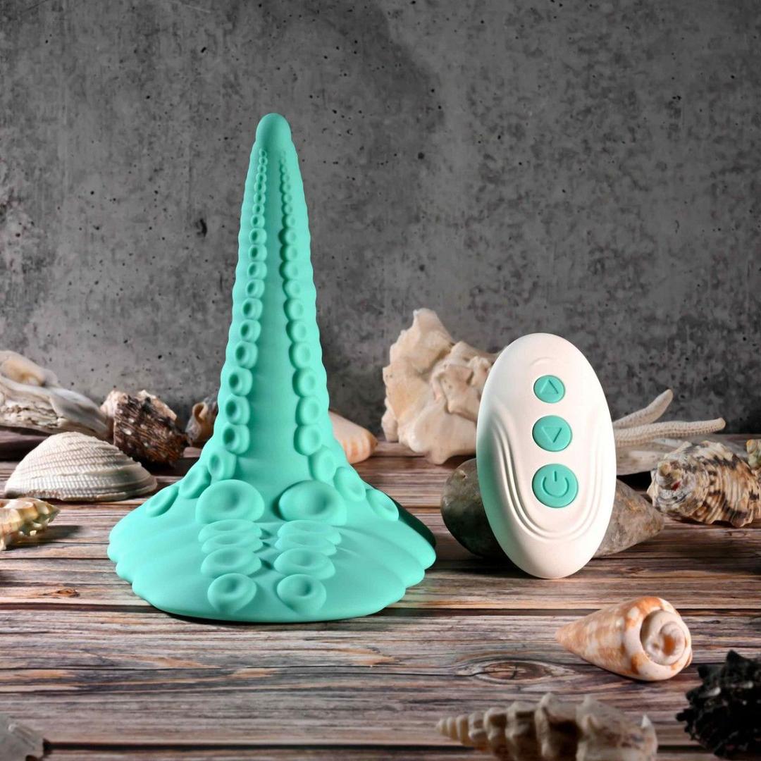 The Evolved Tentacle Grinder by Evolved Novelties, a teal silicone suction-cup toy, stands upright on a wooden surface next to a white remote control, surrounded by seashells, with a gray textured background visible.