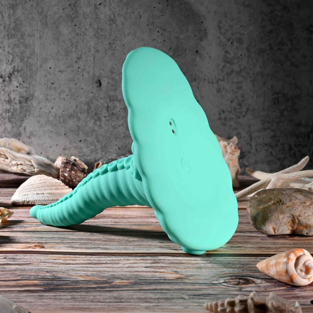 The Evolved Novelties Evolved Tentacle Grinder—a turquoise, cloud-shaped silicone hands-free vibrator with a ribbed handle—stands upright on wood, surrounded by seashells and rocks against a gray textured background.