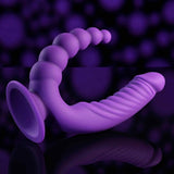 The Evolved Novelties Evolved The Rider is a purple silicone double-ended vibrator with a ribbed, curved shaft and rounded, beaded tip. Waterproof for hands-free, versatile pleasure, it’s shown against a dark background with purple bokeh lights.