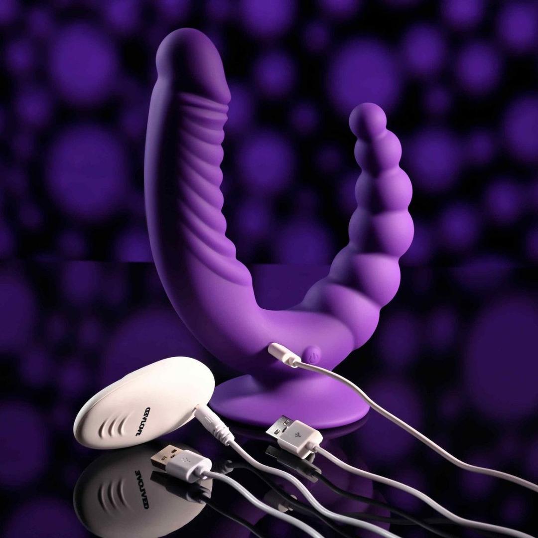 The Evolved Novelties Evolved The Rider, a purple double-ended vibrator with a curved, ribbed design, is shown with white USB charging cables and a remote against a purple and black blurred background.