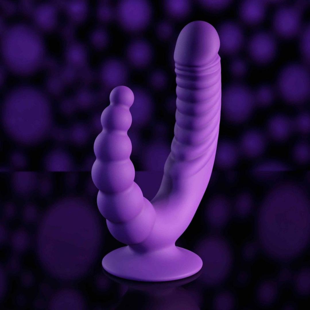 The Evolved The Rider by Evolved Novelties is a purple silicone double-ended vibrator with a ribbed texture and a suction base, displayed on a dark background with subtle, blurred purple lights.
