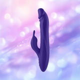 The FemmeFunn Booster Rabbit XL Dark Purple by Femme Funn is a dark purple, curved dual-action vibrator with a smaller extension, crafted from waterproof silicone and set against a soft, blurred background with pink, purple, and blue bokeh lights.