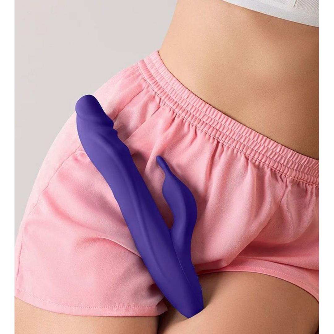 A person in pink shorts and a white top holds the Femme Funn Booster Rabbit XL Dark Purple vibrator by FemmeFunn, made from waterproof silicone, against their shorts near the pelvic area.