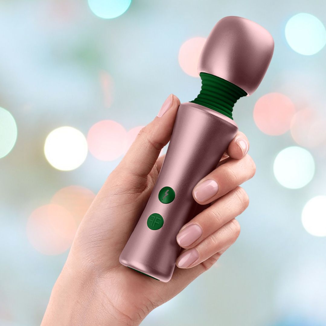 A hand holding a compact, metallic rose gold Femme Funn Bougie Wand Massager with green buttons. The background is softly blurred with various circular pastel lights, highlighting the massager's design and emphasizing its powerful vibrations.