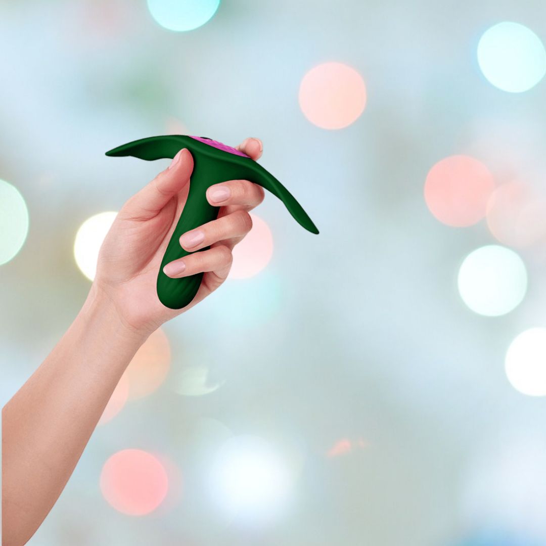 A hand is holding a dark green Femme Funn Ossia Wearable Bullet Vibrator by Femme Funn, featuring a unique curved design. Set against a background of blurred, colorful bokeh lights, the vibrator has an ergonomic shape and appears to have a button on top.