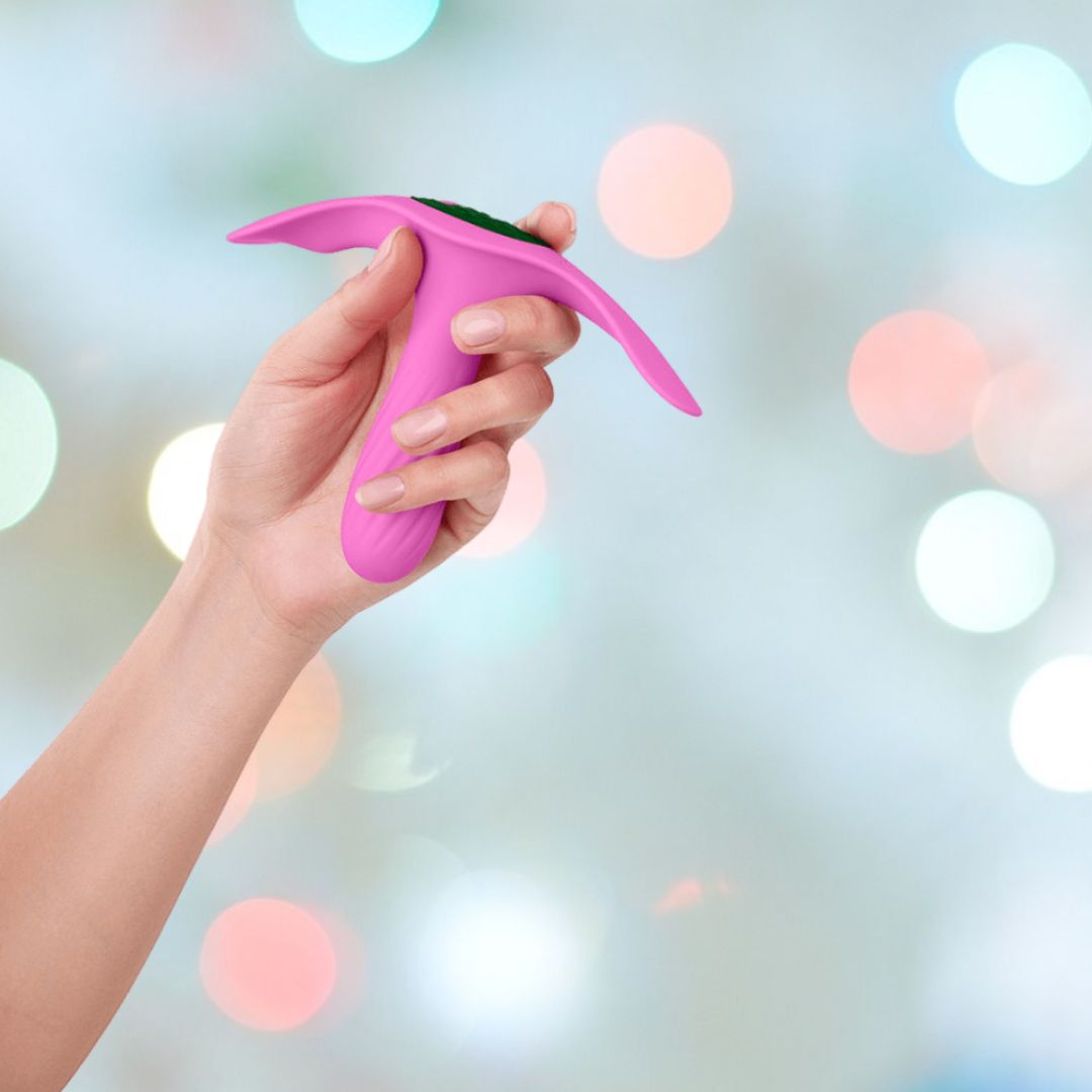 A hand holding a pink Femme Funn Ossia Wearable Bullet Vibrator with a curved top against a soft, out-of-focus background with colorful circular lights.