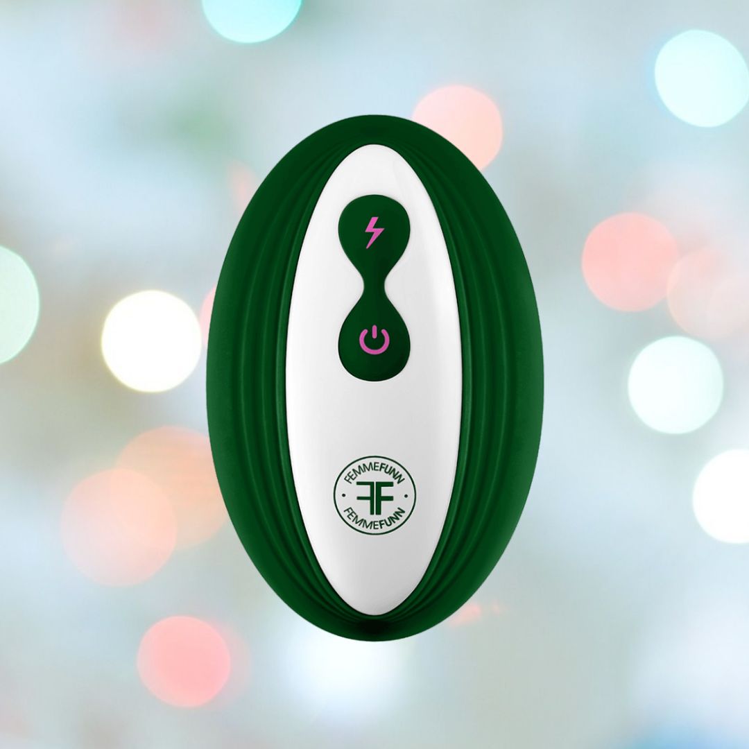 A wearable bullet vibrator named the Femme Funn Ossia by Femme Funn, features a dark green outer case and white center with a pink lightning bolt icon above the power button. The background is out-of-focus with soft, colorful circular lights, and it has a round logo with "TF" along with some text near the bottom, resembling a handheld electronic device.