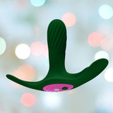 A Femme Funn Ossia Wearable Bullet Vibrator in dark green, featuring a three-pronged design with a textured surface and a pink circular center section, is displayed against a soft, blurred background adorned with pastel-colored bokeh lights.