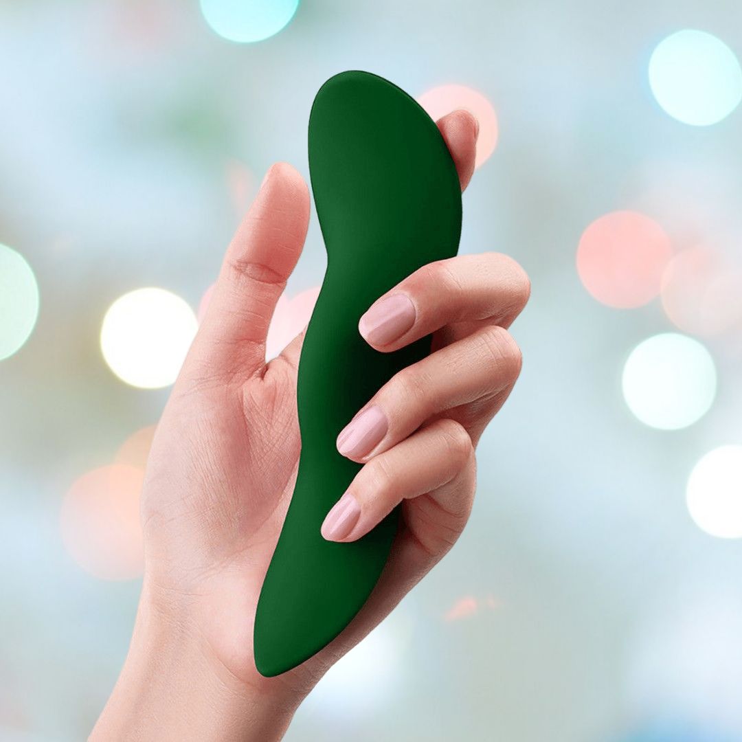 A hand with neatly manicured nails holds a sleek, ergonomically designed FemmeFunn Unda Panty Vibrator Dark Green against a blurred background of colorful lights. The device boasts a smooth, contoured shape for discreet pleasure and even includes a Boost Mode for an enhanced experience.