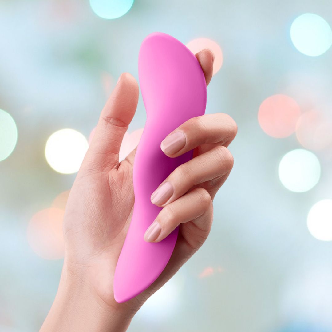 A hand with neatly manicured nails holds a pink, curved, and smooth object against a blurred background with colorful bokeh lights. The ergonomic design of the FemmeFunn Unda Panty Vibrator Pink ensures it fits comfortably in your hand.